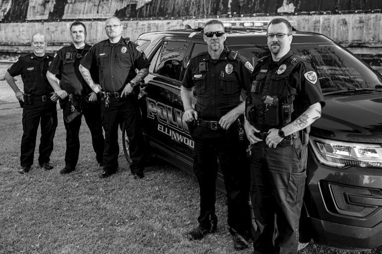 Police officers standing beside a City of Ellinwood Police car. 2 officers are at the front of the vehicle and 3 officers are standing near the rear of the vehicle.