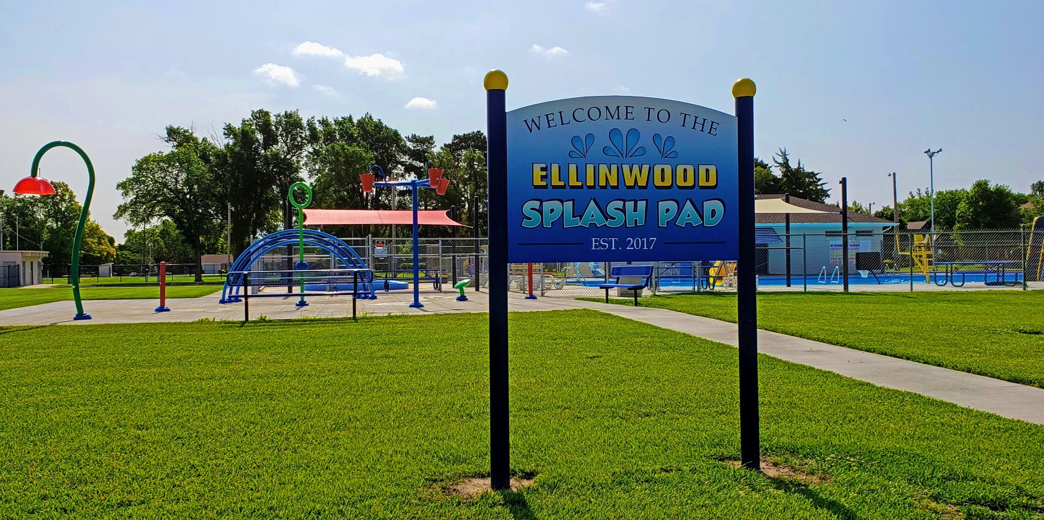 A sign in the foreground reads, "Welcome to the Ellinwood Splash Pad. Established 2017". A splash pad and pool can be seen in the background.
